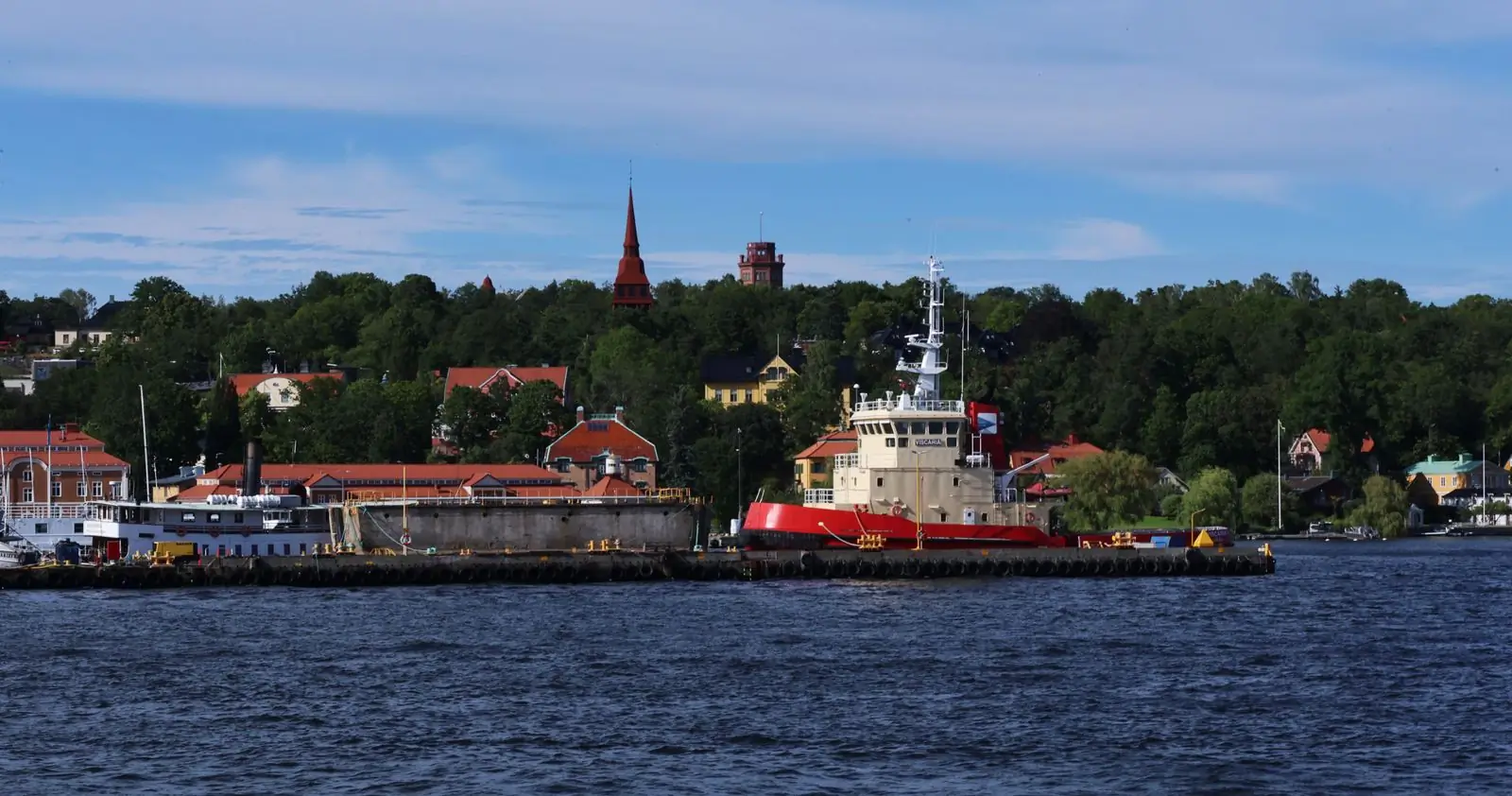 Bogserbåt vid kaj i Stockholm, med hus och ett kyrktorn bland träden bakom